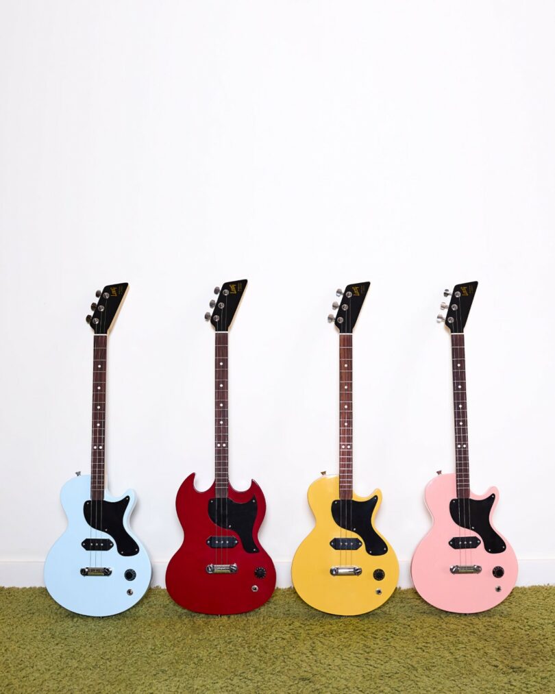 Four electric guitars in blue, red, yellow, and pink stand upright on a green carpet against a plain white wall—perfect inspiration for any Design Milk gift guide.