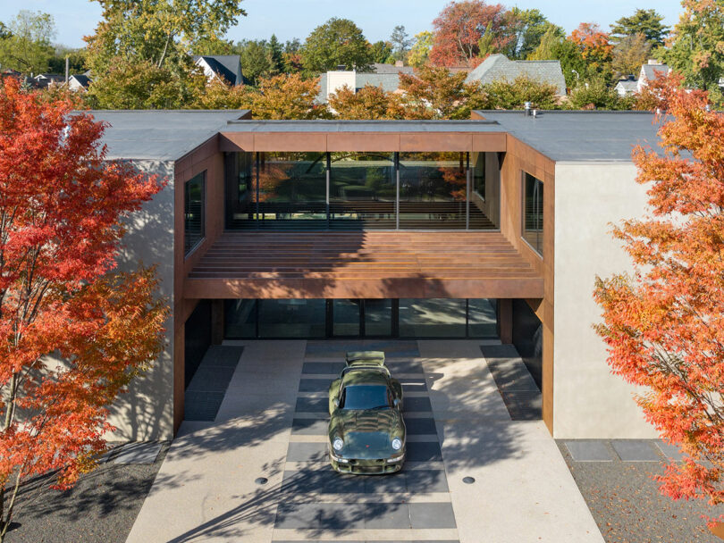 A green vintage car is parked in the driveway of a modern, two-story house with large windows, surrounded by trees with autumn-colored leaves.