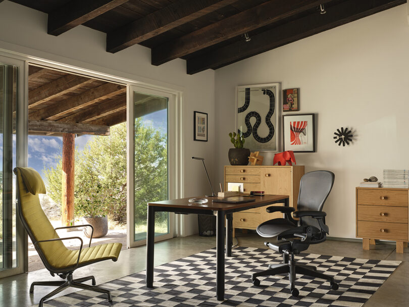 Modern home office with wooden ceiling beams, large glass doors, a black office chair at a desk, a yellow lounge chair, and art on the walls, set on a checkered rug.