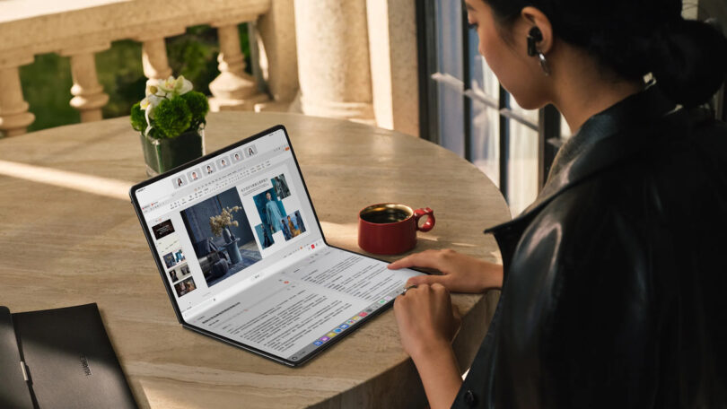 A person explores the latest technology on a foldable tablet with a large screen at a round table, beside a red coffee cup and a small potted plant.