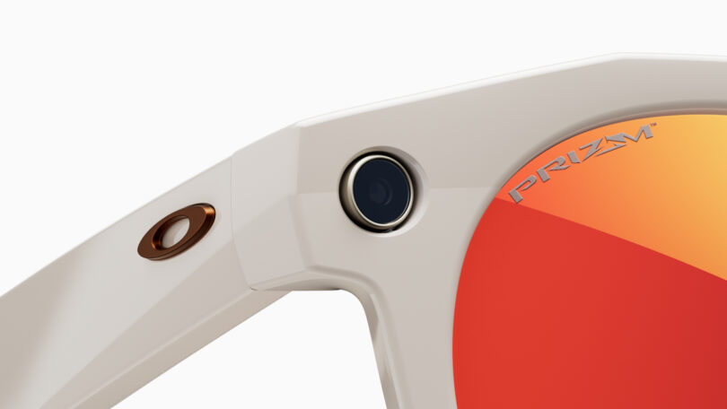 Close-up of the upper corner of beige sunglasses with an orange reflective lens, showcasing innovative technology—a small built-in camera on the frame—and the word “PRIZM” printed on the lens.