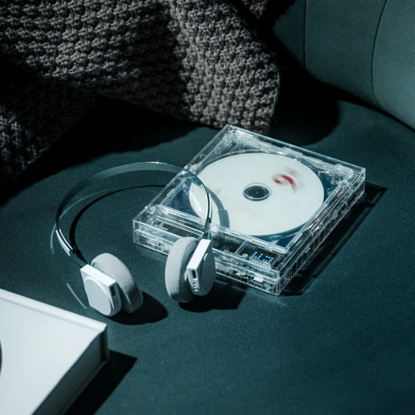 A pair of modern headphones and a transparent CD player with a disc inside showcase sleek technology as they rest on a dark green fabric surface next to a textured blanket.
