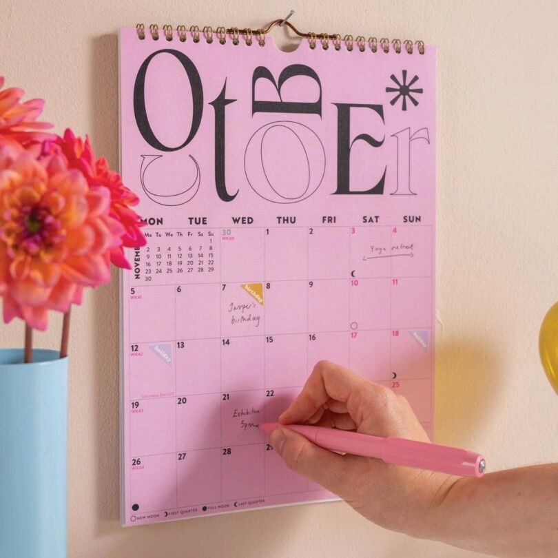 A hand writes on a pink 2026 calendar for October, which hangs next to a blue vase with orange flowers.
