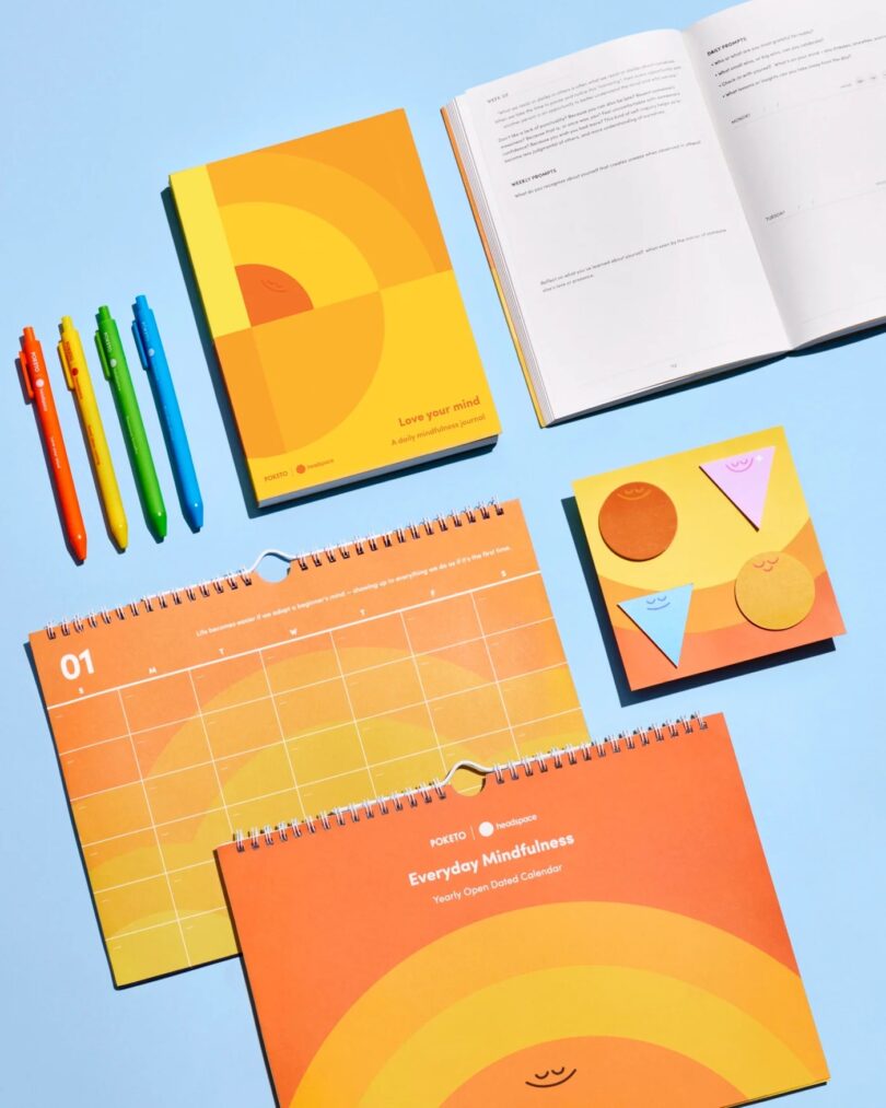 An assortment of stationery on a blue surface, including 2026 calendars, a journal, colorful pens, sticky notes, and an open notebook.