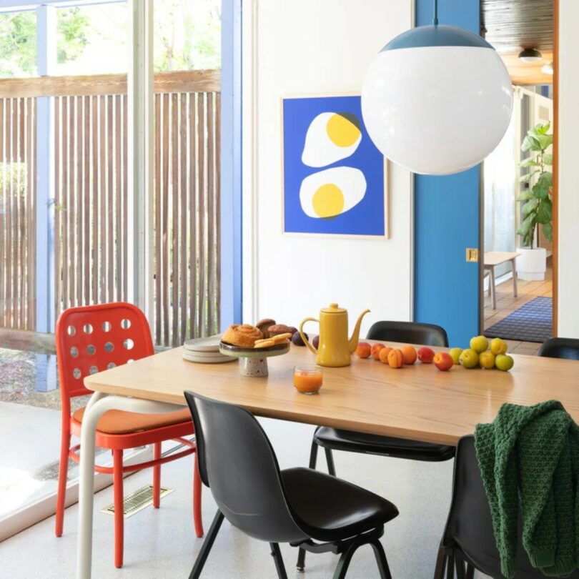 A dining table with black and red chairs, a yellow teapot, pastries, fruit, and juice. A large round pendant light hangs above; abstract art and windows are in the background.