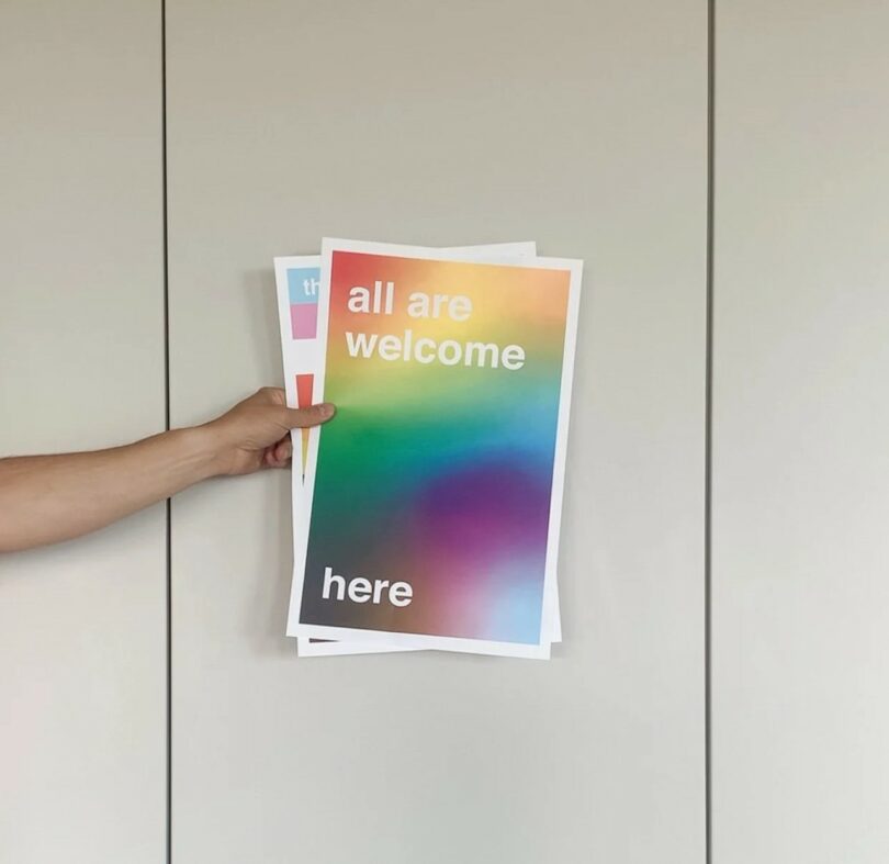 A hand holds posters, with the top one displaying a rainbow gradient background and the text "all are welcome here