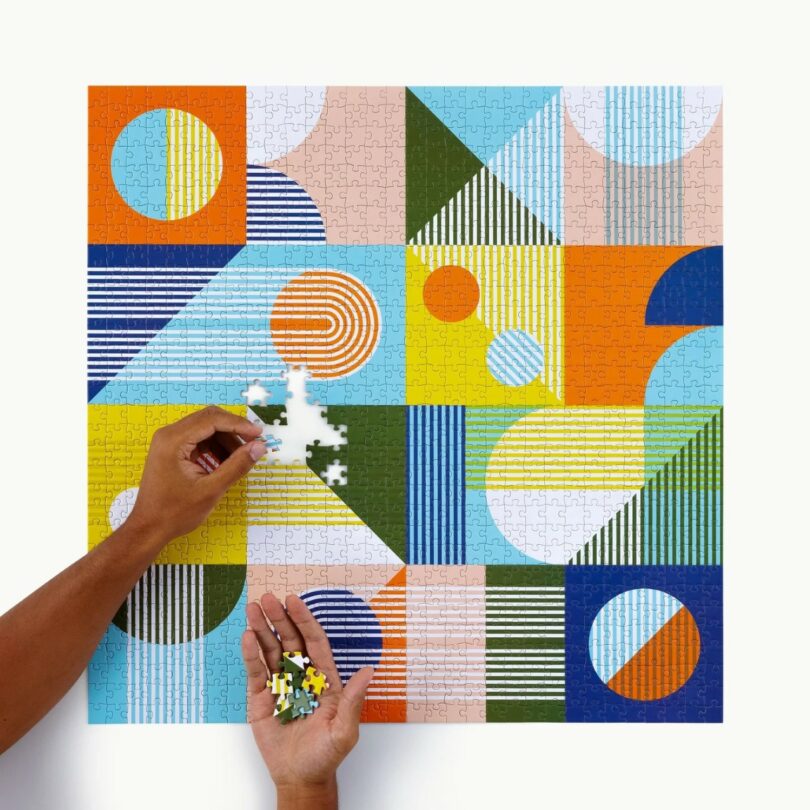 Two hands work on a colorful geometric jigsaw puzzle, holding and placing pieces against a mostly completed puzzle background with various shapes and patterns