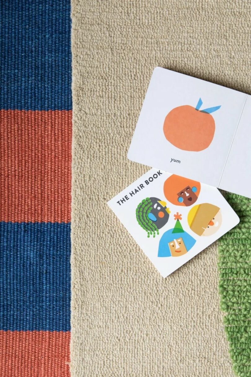 Two children's board books are open on a textured rug; one shows an orange fruit, the other has illustrated faces with different hairstyles