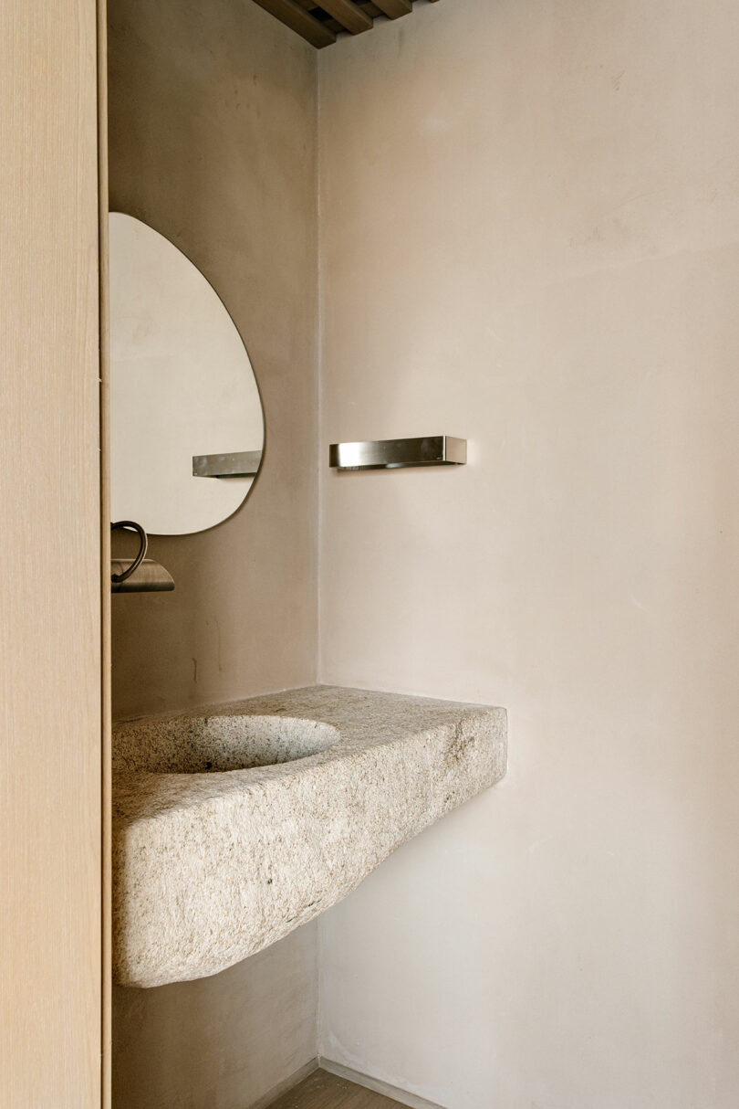 Minimalist bathroom with a stone sink, an oval mirror, a metallic towel bar, and beige plaster walls.