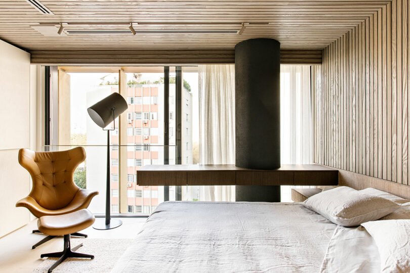 Modern bedroom with wood-paneled walls and ceiling, large bed, tan lounge chair, floor lamp, and floor-to-ceiling windows overlooking an apartment building.
