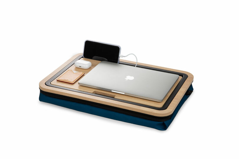 Portable lap desk with a closed laptop, a smartphone being charged, wireless earbuds case, and a small wallet placed on a wooden surface with a blue cushion underneath.