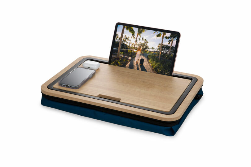 A tablet displaying a tropical scene and a smartphone with earbuds rest on a wooden lap desk with a blue cushion base.