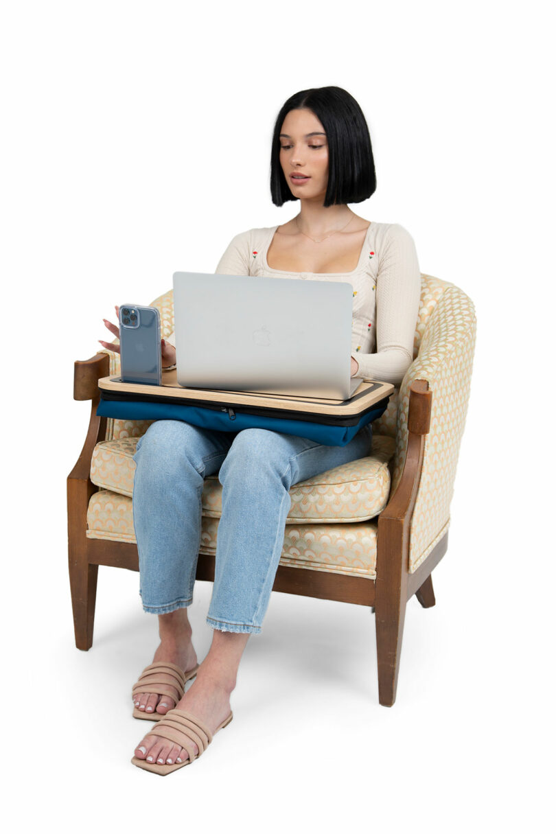 Woman sitting in an upholstered chair using a laptop on a lap desk, with a smartphone placed upright in a holder on the desk.