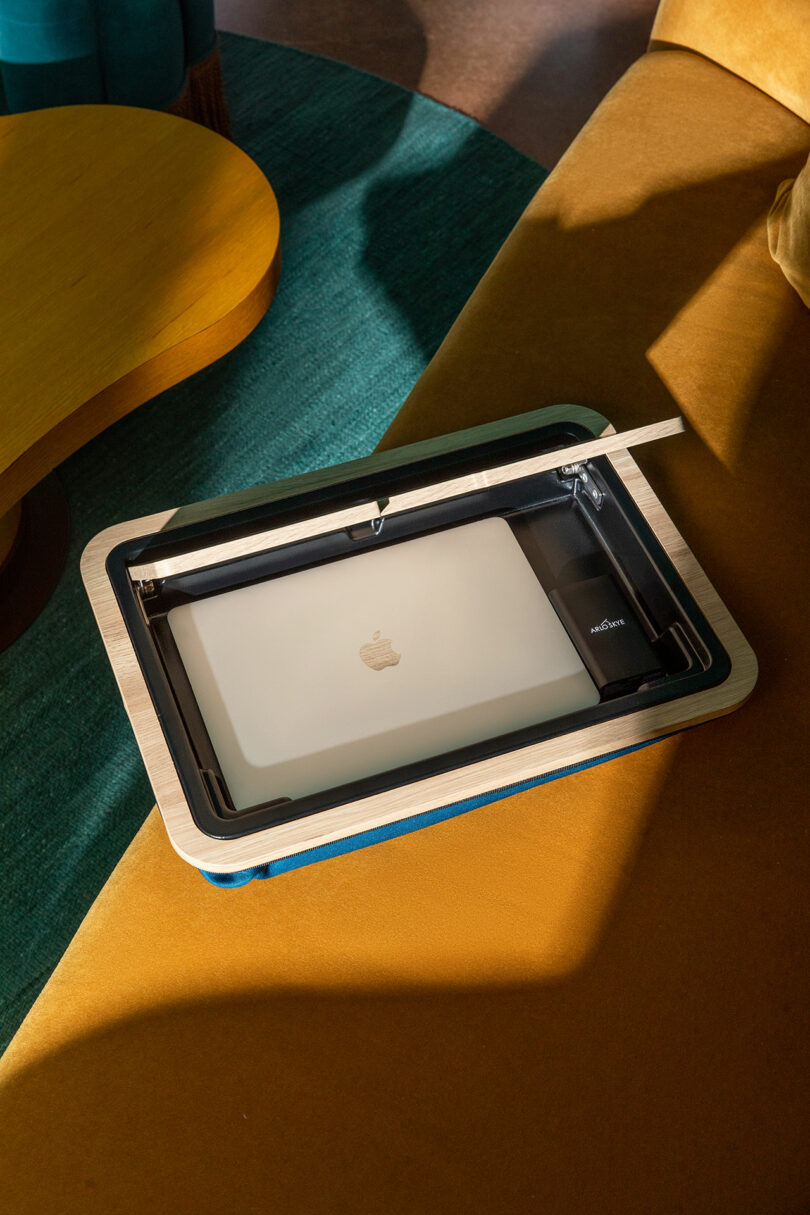 A closed silver MacBook, a black water bottle, and a pen are placed in a black tray on a mustard yellow couch, next to a teal rug and a wooden table.