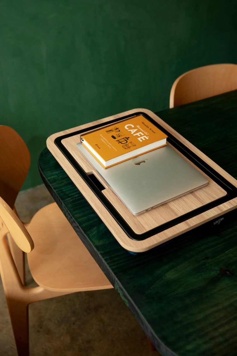 A closed laptop and a yellow book are stacked on a wooden tray placed on a dark green table next to a wooden chair.