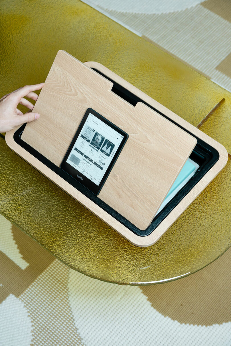 A hand slides open a wooden desk organizer on a glass table, revealing an e-reader and some papers stored inside.