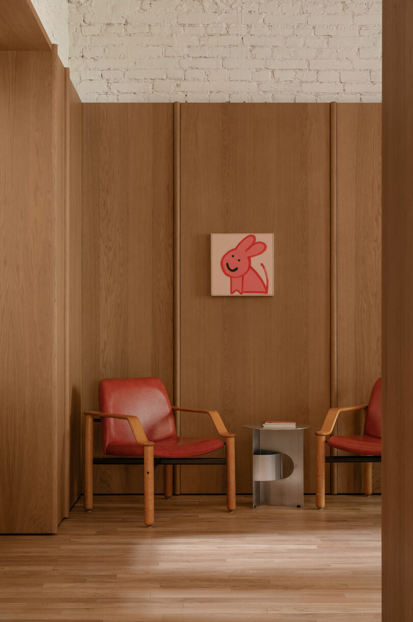 Two red chairs with wooden arms flank a small clear side table in a room with wood paneling and a white brick wall. A pink artwork featuring a cartoon rabbit hangs above.