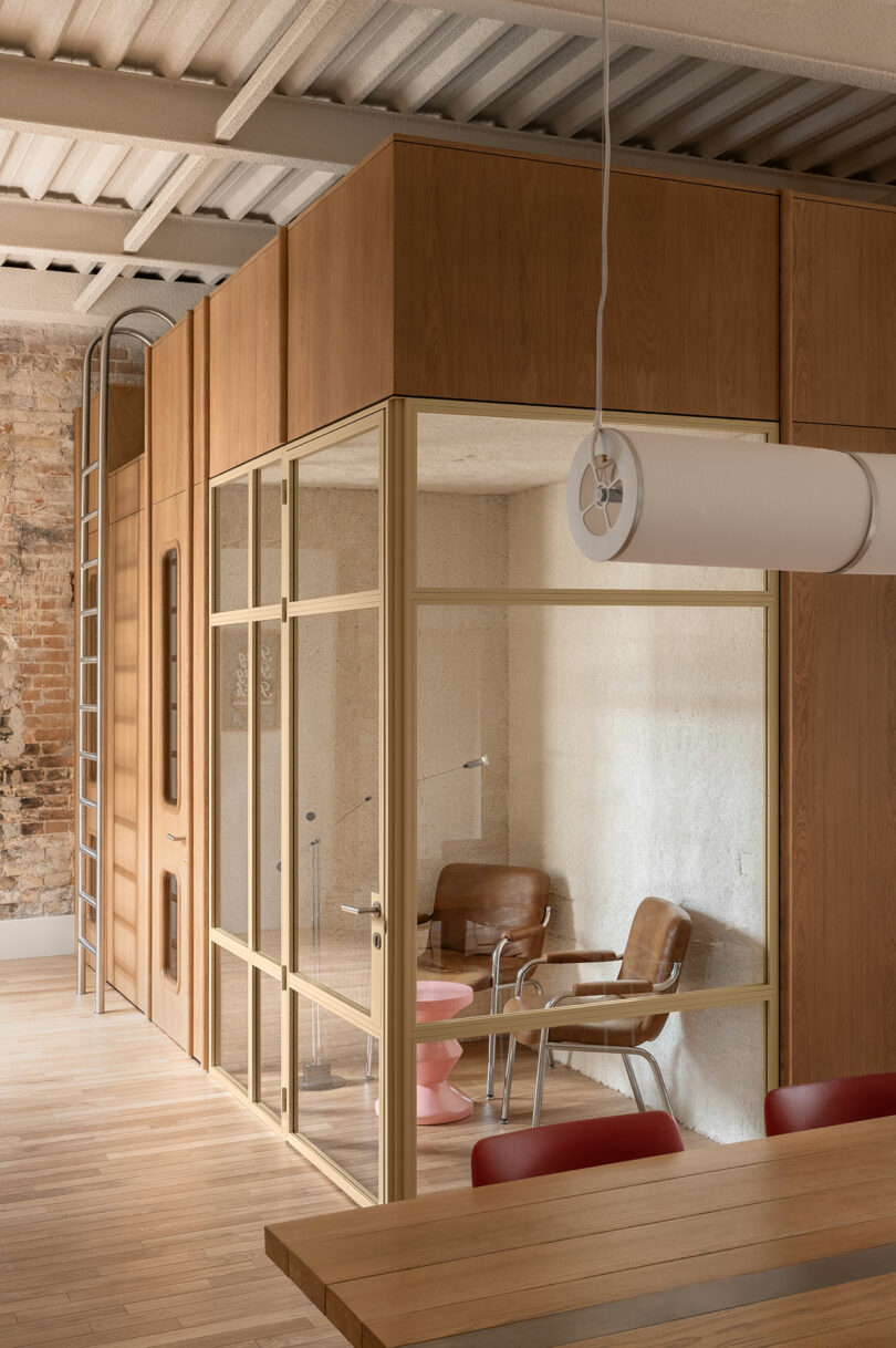 A small office room with glass walls, two brown leather chairs, a pink side table, and a metal ladder inside a modern space with wood accents and an exposed brick wall.