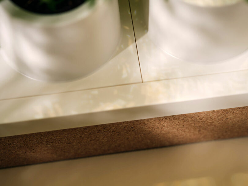 Two white ceramic pots are placed on a white surface next to a brown cork board, with light and shadow patterns visible on the surface.