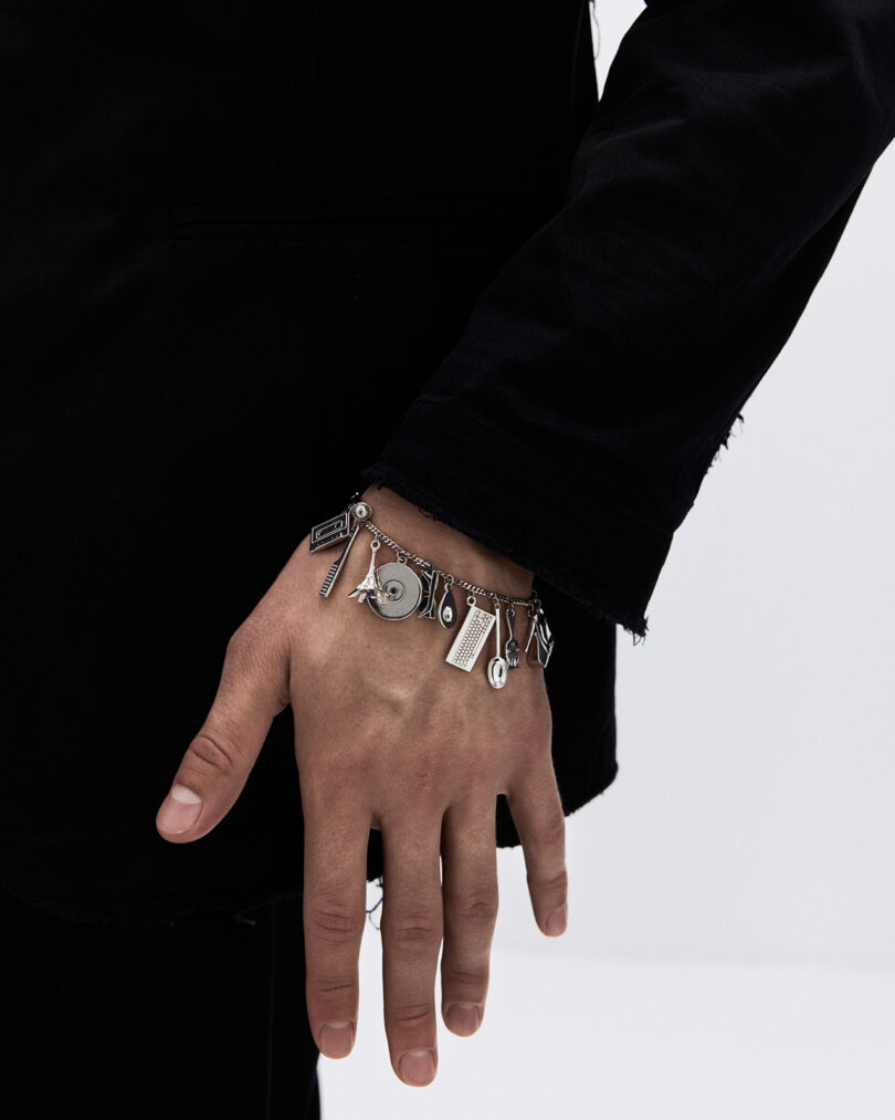 A person wearing a dark jacket displays a silver charm bracelet on their left wrist, with various small charms hanging from the chain.
