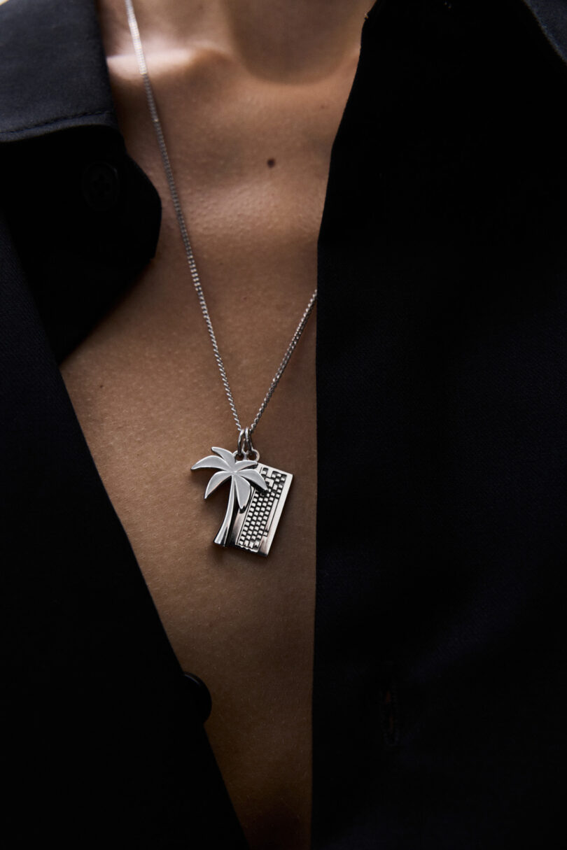 A person wearing a black shirt with the top buttons undone displays a silver necklace featuring a palm tree and keyboard pendant.