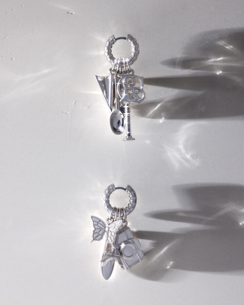 Two silver hoop earrings, each with multiple metallic charms, are displayed on a light gray background with shadows and reflections.