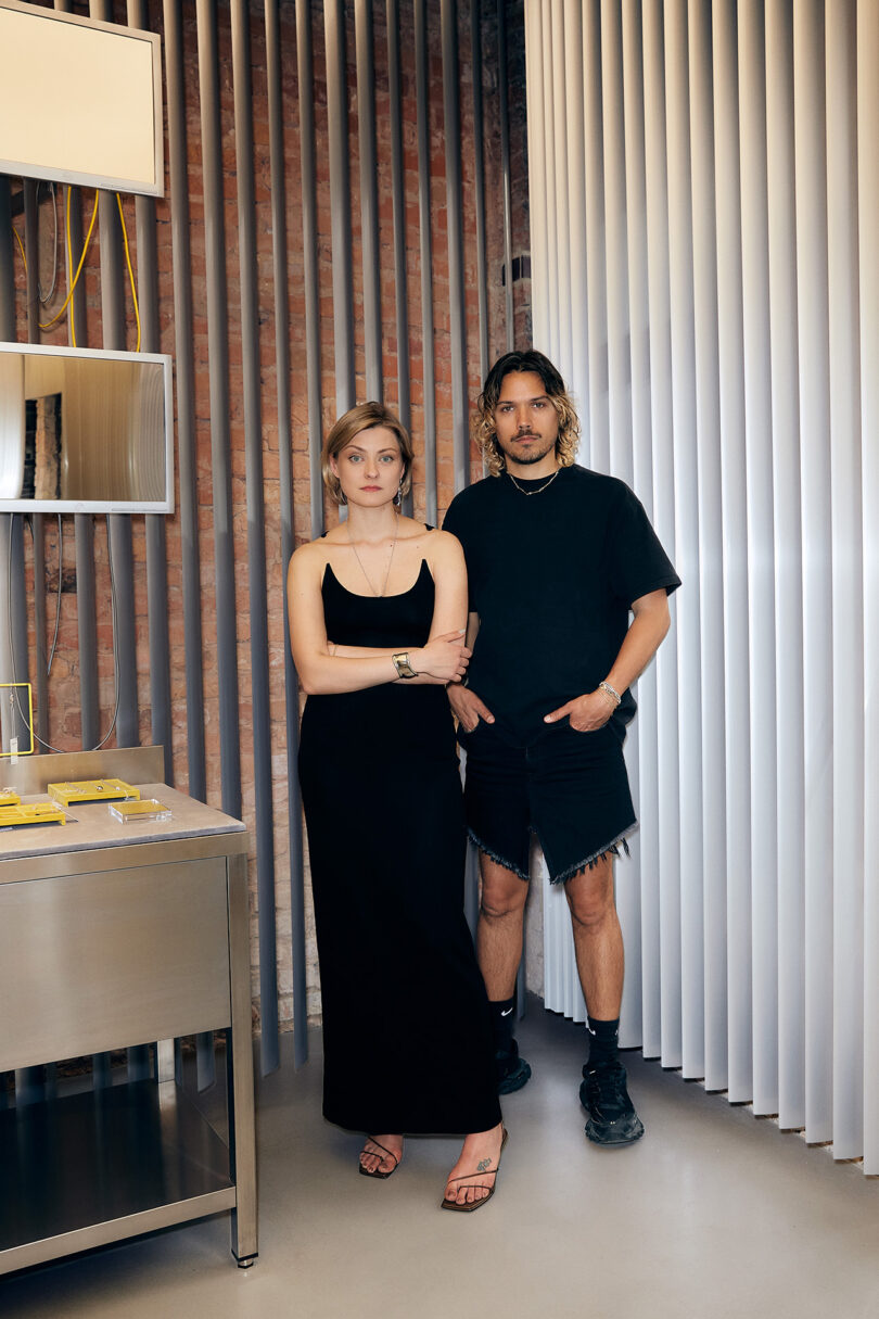 Two people stand side by side indoors; the woman wears a long black dress and sandals, the man wears a black t-shirt, shorts, and sneakers. Vertical blinds and brick wall are in the background.
