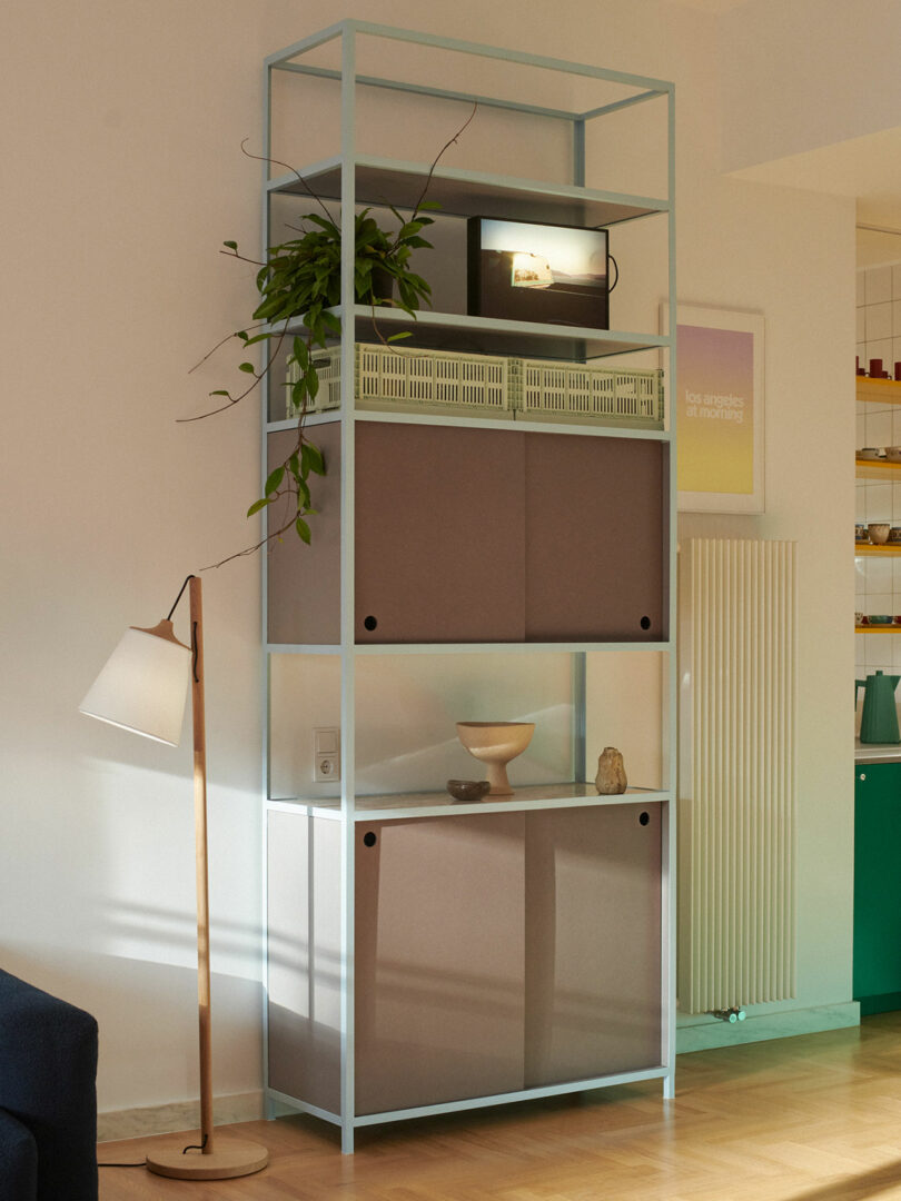 A tall, light blue metal shelving unit with plants, a lamp, boxes, and decorative items stands against a wall in a modern living room.