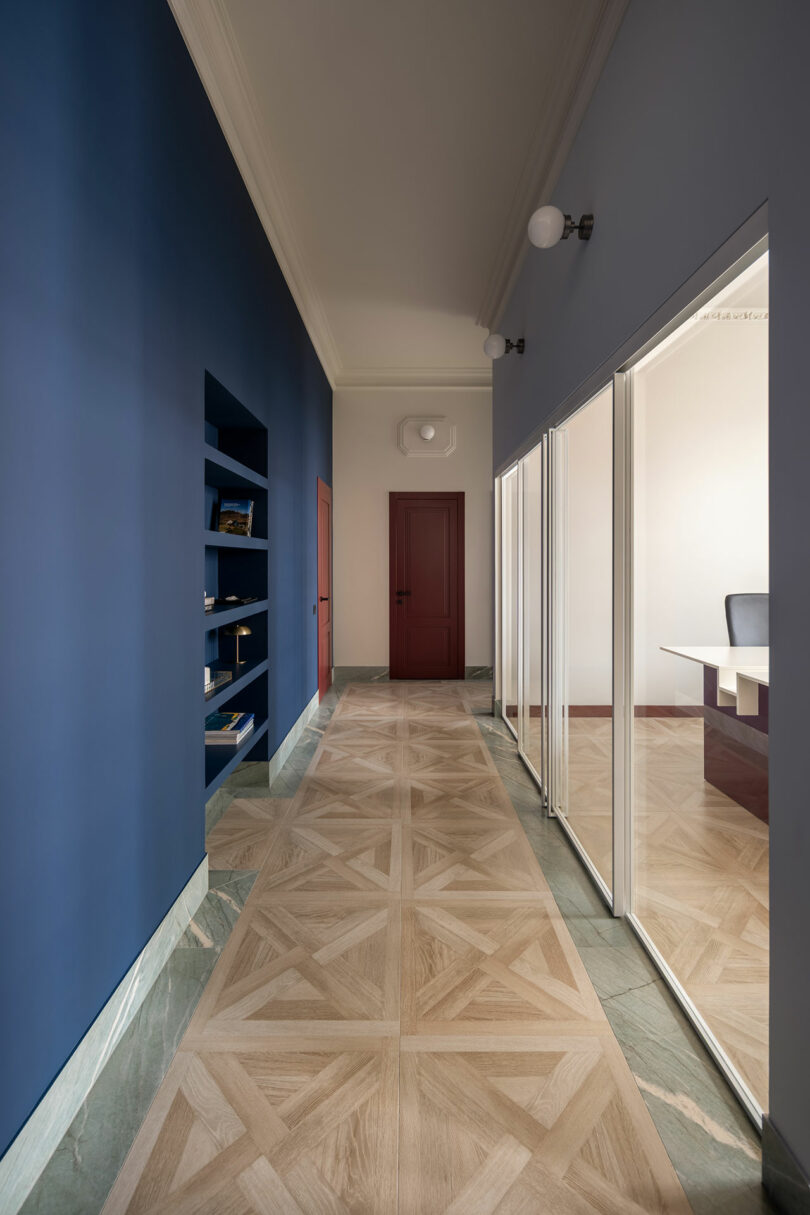 A hallway with blue walls, built-in shelves, patterned wood flooring, glass office partitions on the right, and a red door at the end.