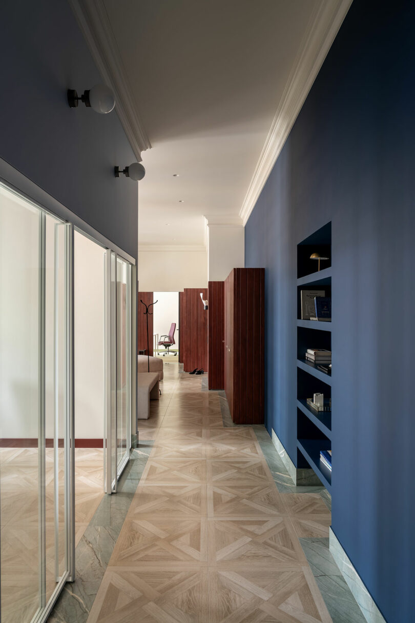 A modern hallway with blue walls, built-in shelves, glass doors on the left, wood flooring, and a view into a living area with wooden furniture.