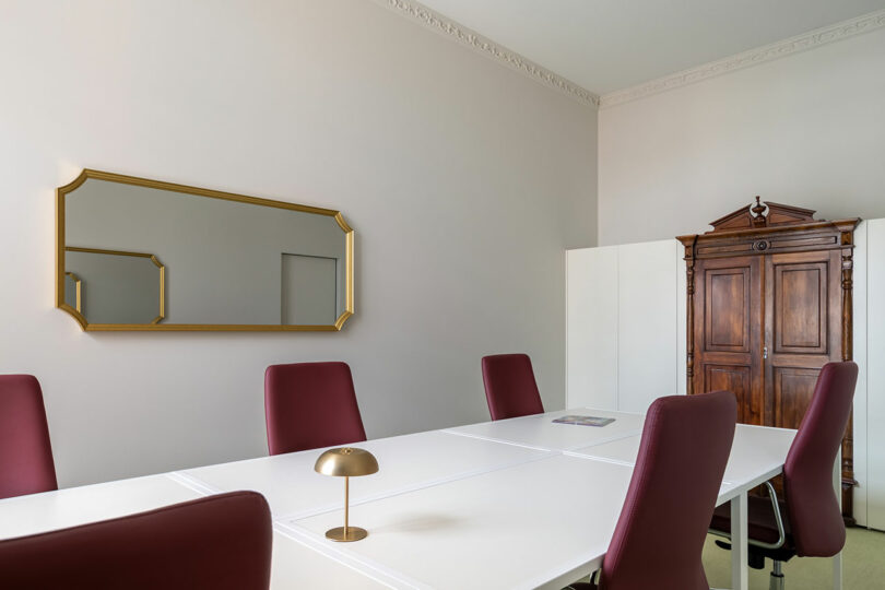 A conference room with a white table, six maroon chairs, a gold-framed mirror, a small lamp, and a large wooden cabinet against a white wall.