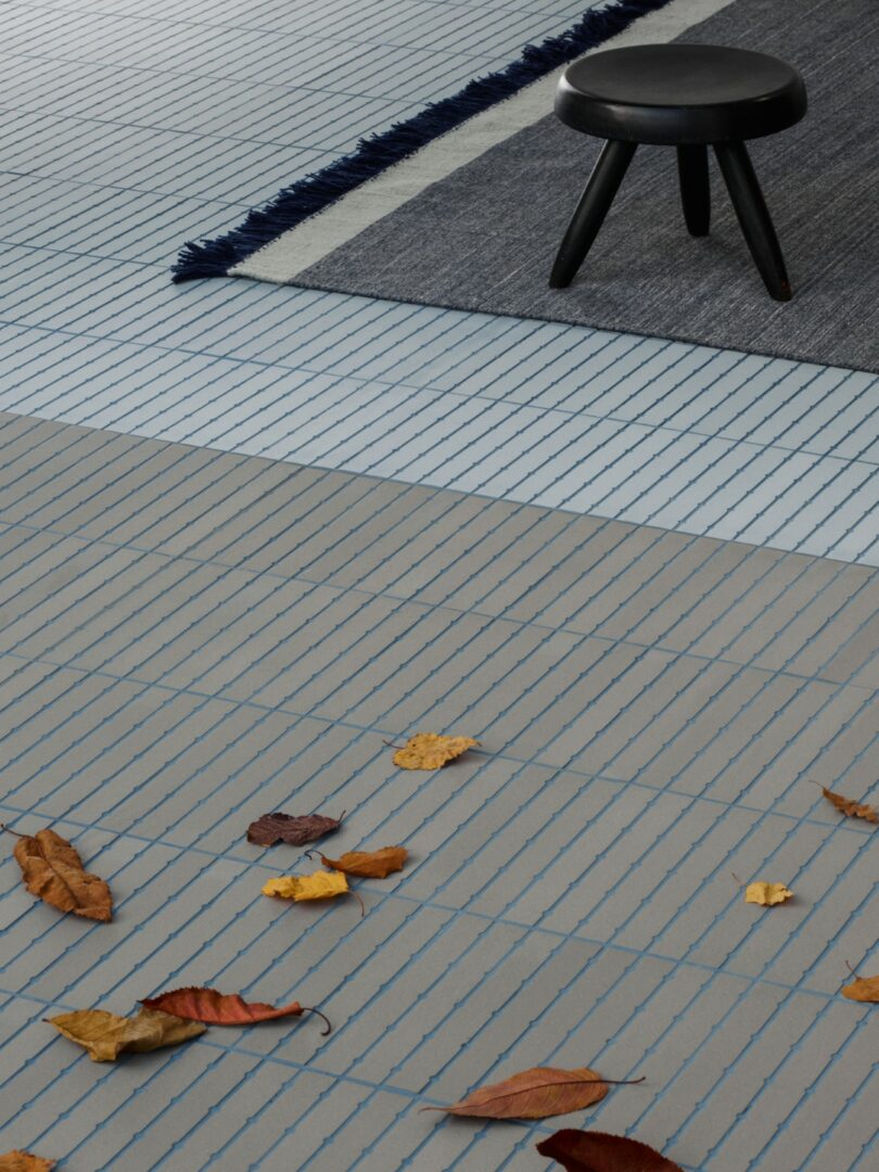 A black stool on a gray rug with fringed edges, placed on a tiled floor scattered with brown leaves