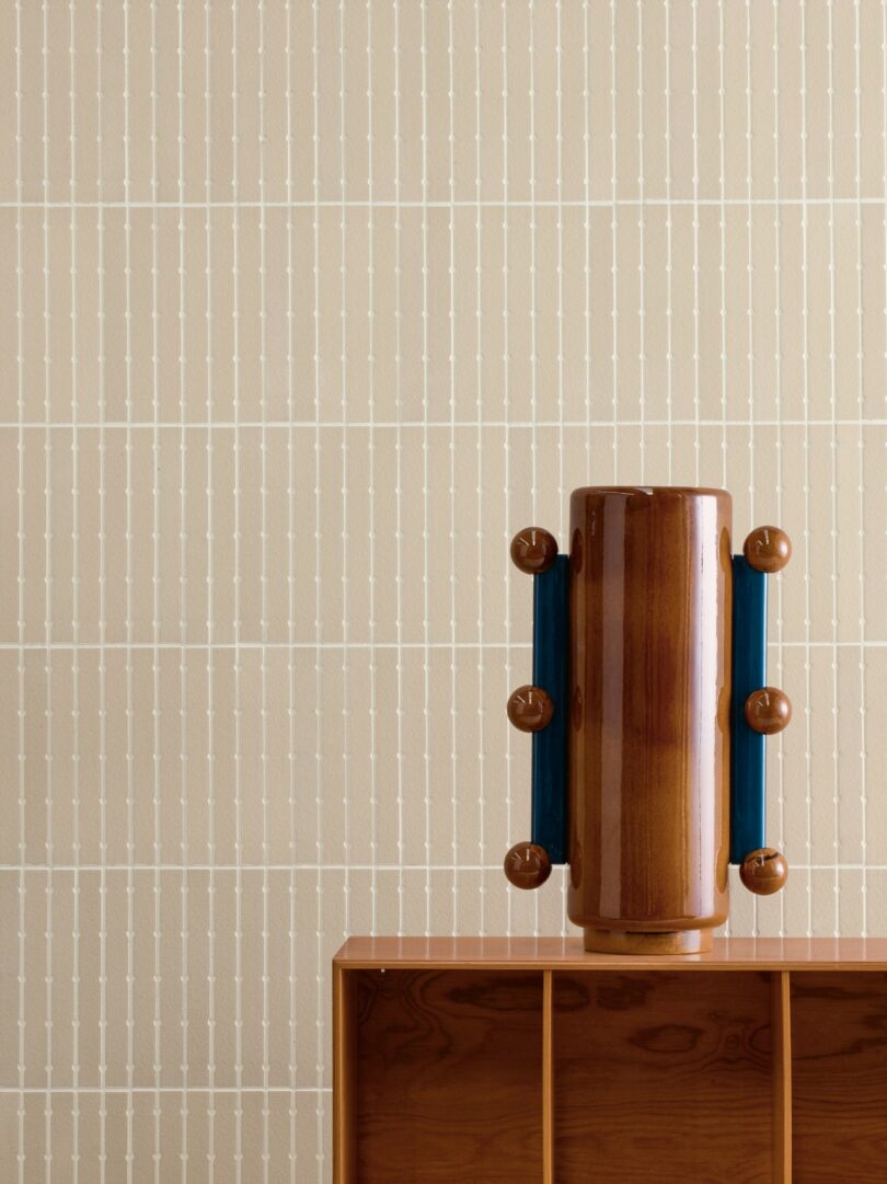 A cylindrical wooden vase with blue side handles sits on a wooden shelf against a beige patterned wall