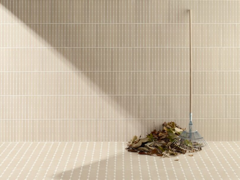 A pile of dry leaves is gathered against a tiled wall, with a blue rake leaning upright next to it on a patterned floor