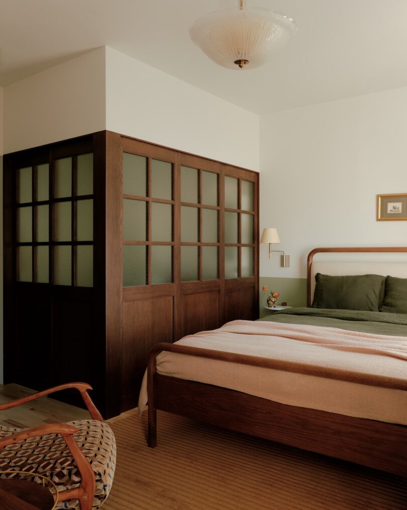 Bedroom with a wooden bed, green bedding, a woven chair, and a dark wood partition with frosted glass panels separating the bed area from the adjacent space.