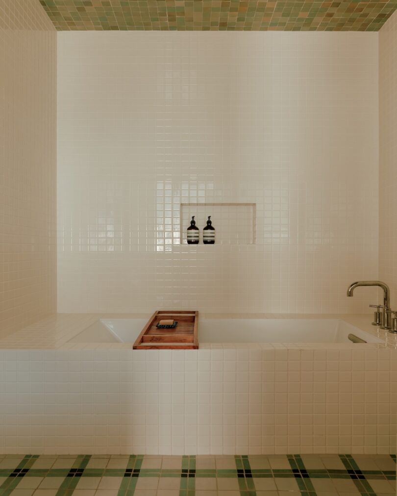 A white tiled bathtub with a wooden tray, two bottles in a recessed wall niche, and green accent tiles on the ceiling and floor.