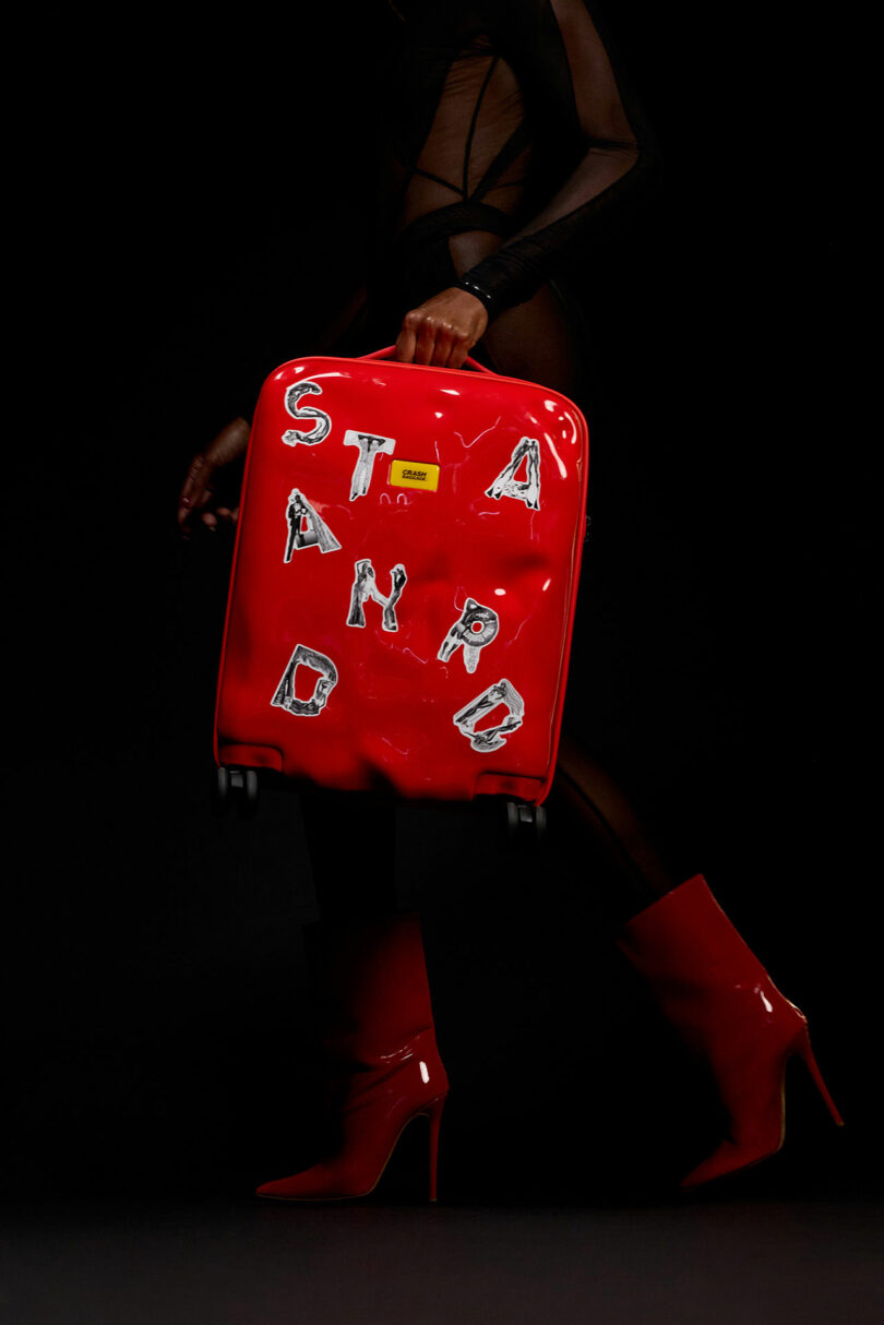 Person in red high-heeled boots and a black sheer outfit holds a red Crash Baggage suitcase with uneven white letters spelling “STANDARD” against a black background.