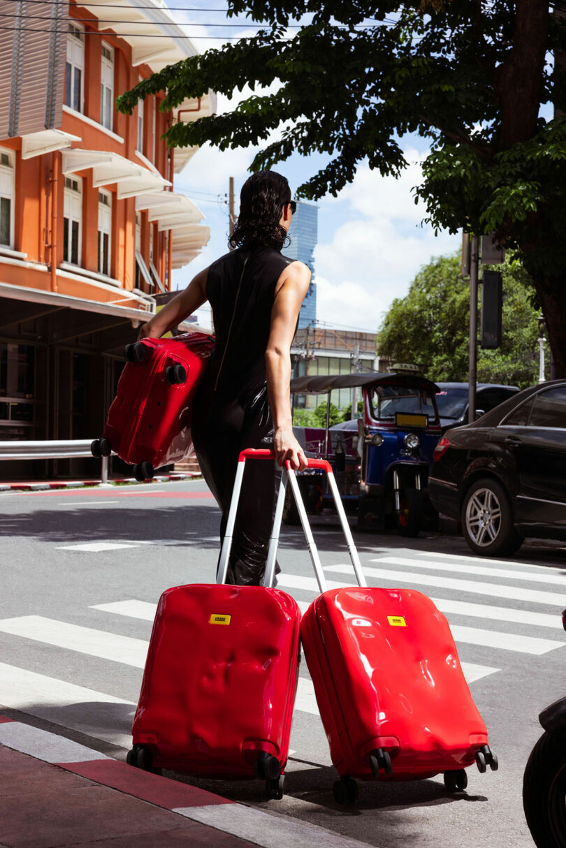A person wearing black clothes crosses a street, pulling two large red Crash Baggage suitcases and carrying a third red suitcase.