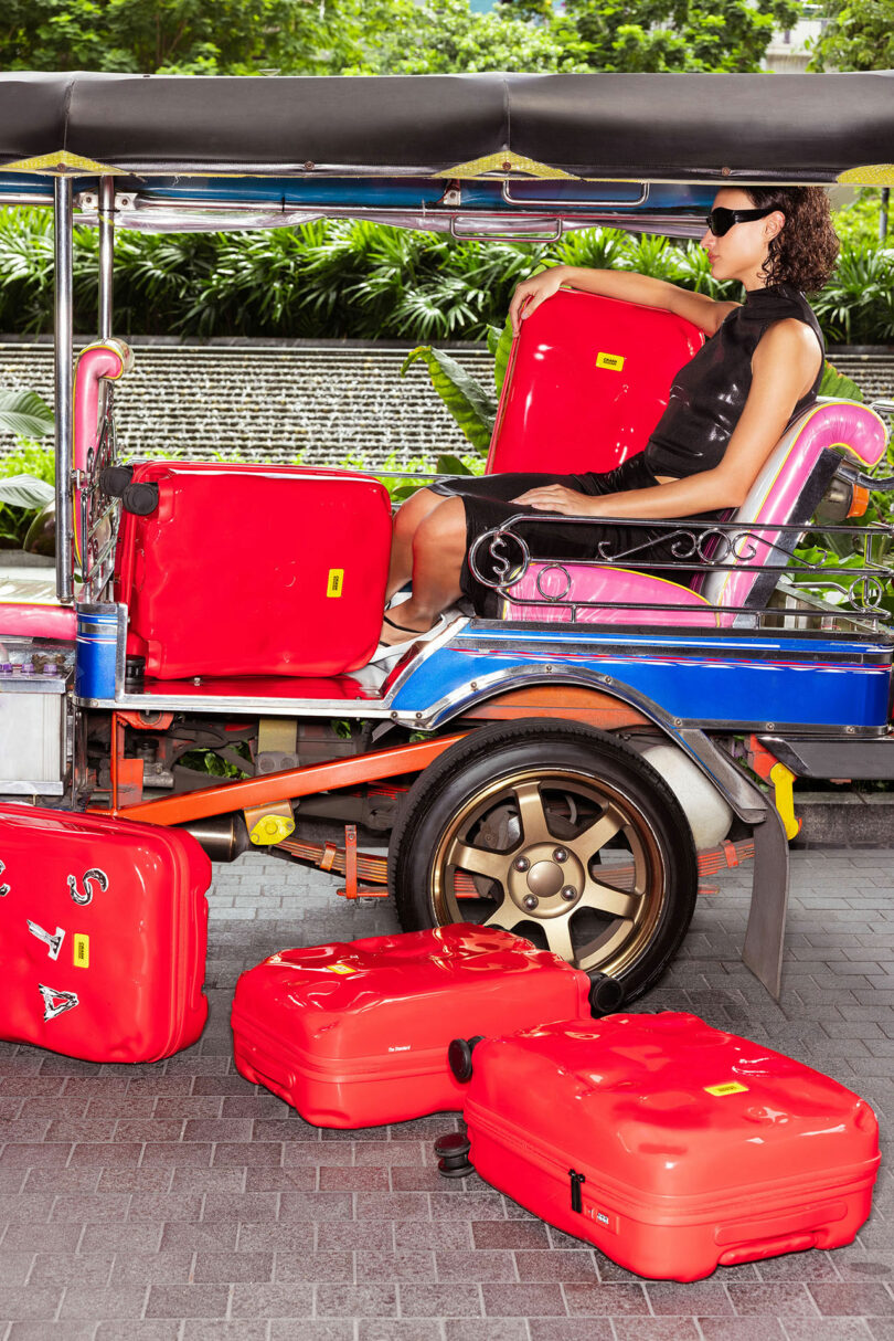 A person wearing sunglasses sits in a tuk-tuk with several bright red Crash Baggage suitcases, some inside the vehicle and others on the ground nearby.