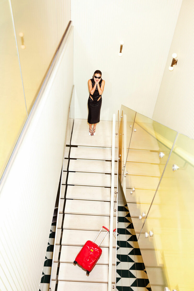 A woman in a black dress stands at the top of a staircase, covering her face, while a red Crash Baggage suitcase lies on the stairs below her.