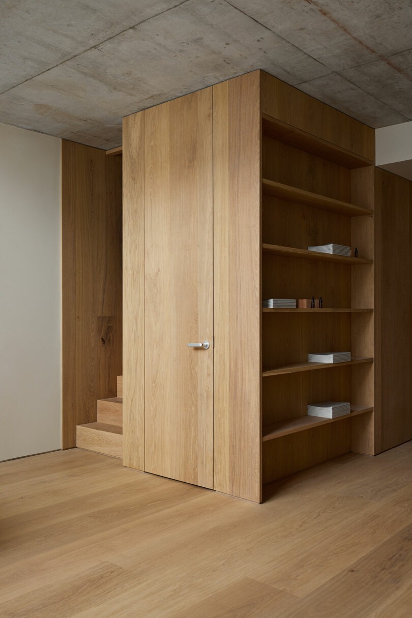 Minimalist interior with wood paneling, built-in shelves holding a few small objects, a closed door, and stairs partially visible in the background.