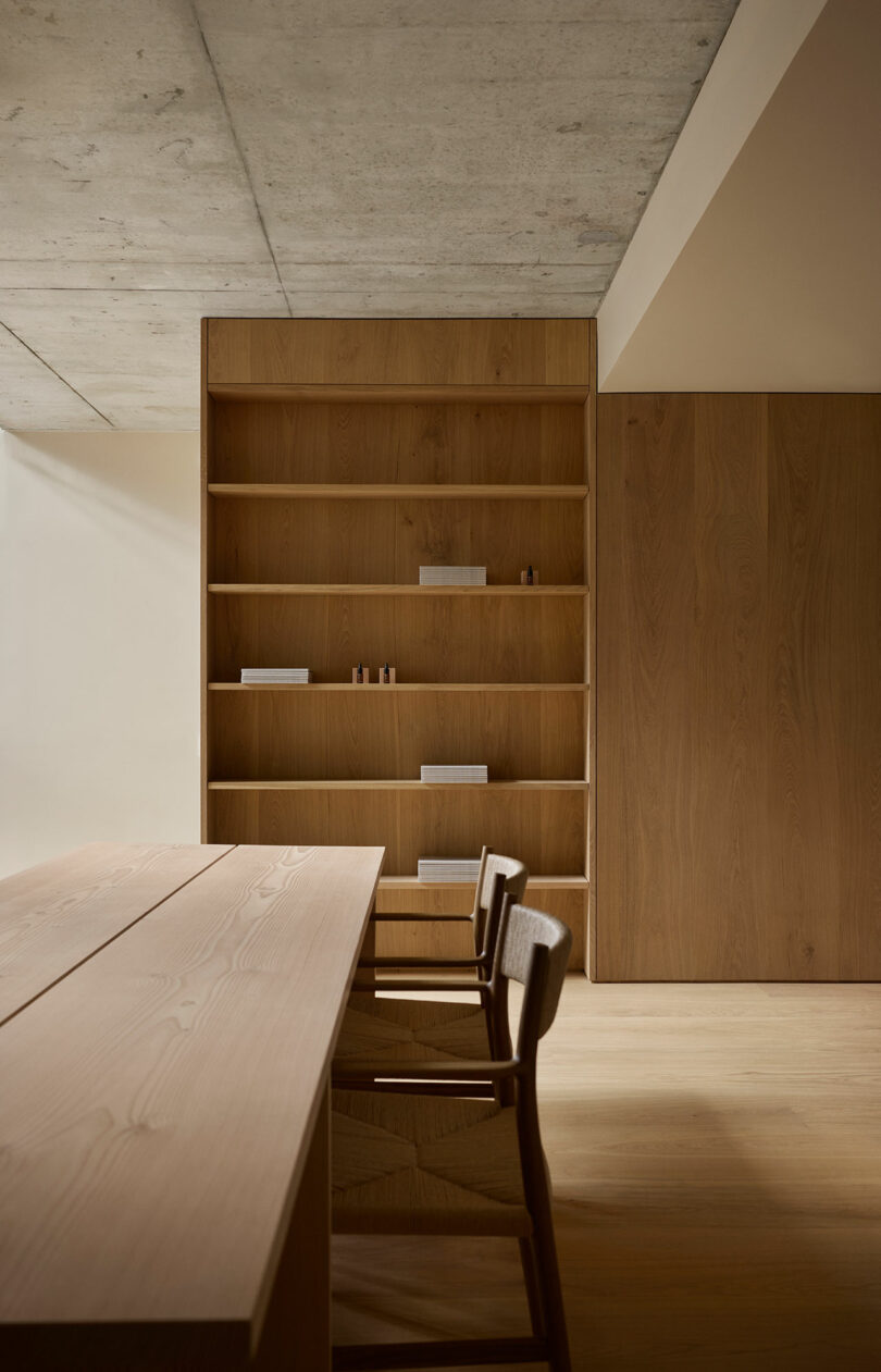 Minimalist room with a wooden table and chairs, and a large wooden bookshelf containing a few small items and books against a light wall.