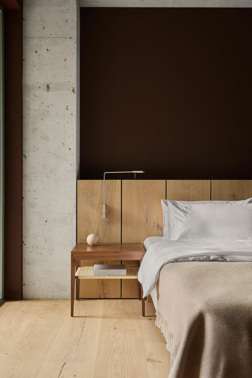A modern bedroom features a wooden bed with white bedding, a beige blanket, a wooden nightstand with a lamp, and a dark brown accent wall beside a concrete pillar.