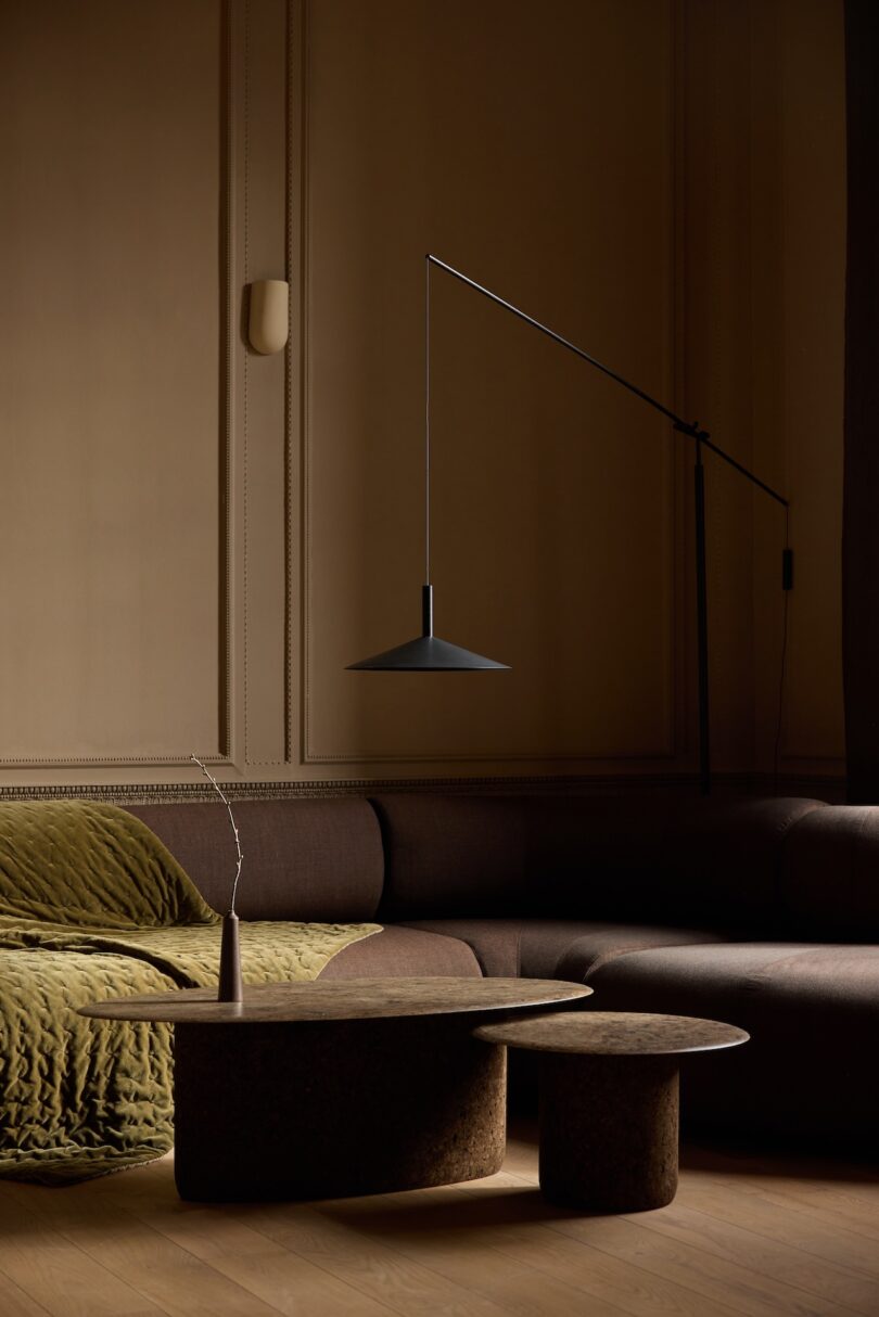 A modern living room with a sectional sofa, round nested coffee tables, a green quilted throw, and a minimalist wall-mounted lamp in soft, warm lighting.