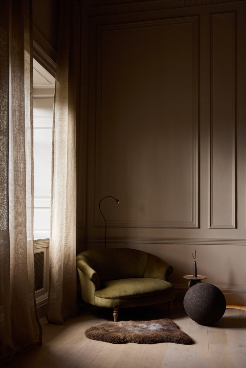 A small green couch sits in the corner of a dimly lit room with tall curtains, a floor lamp, a round black ottoman, and a small side table on a light wooden floor.