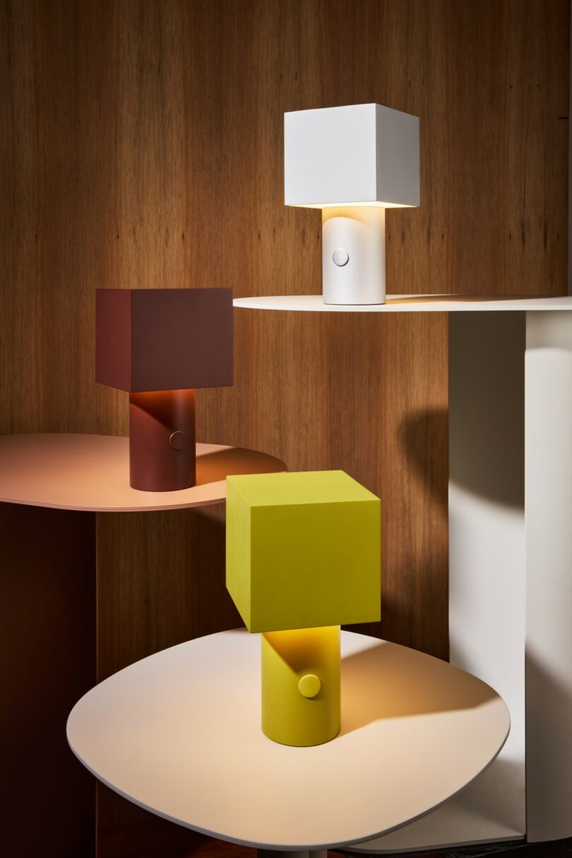 Three modern table lamps with square shades in white, red, and yellow sit on round tables against a wooden wall background