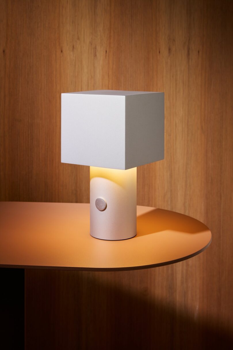 A modern table lamp with a square white shade and cylindrical base sits on a wooden surface against a wood-paneled background