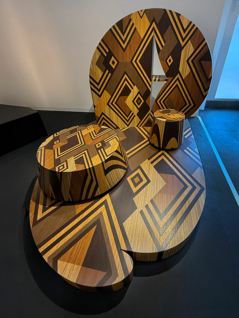 Abstract wooden furniture with geometric, multicolored patterns, featuring a rounded bench, stool, and circular table on a smooth, dark floor.