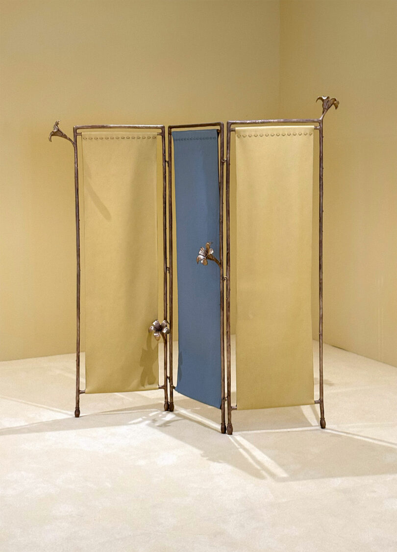 A three-panel folding screen with a metal frame; two beige panels and one blue panel, each with decorative bird accents on the frame, stands in a beige room.