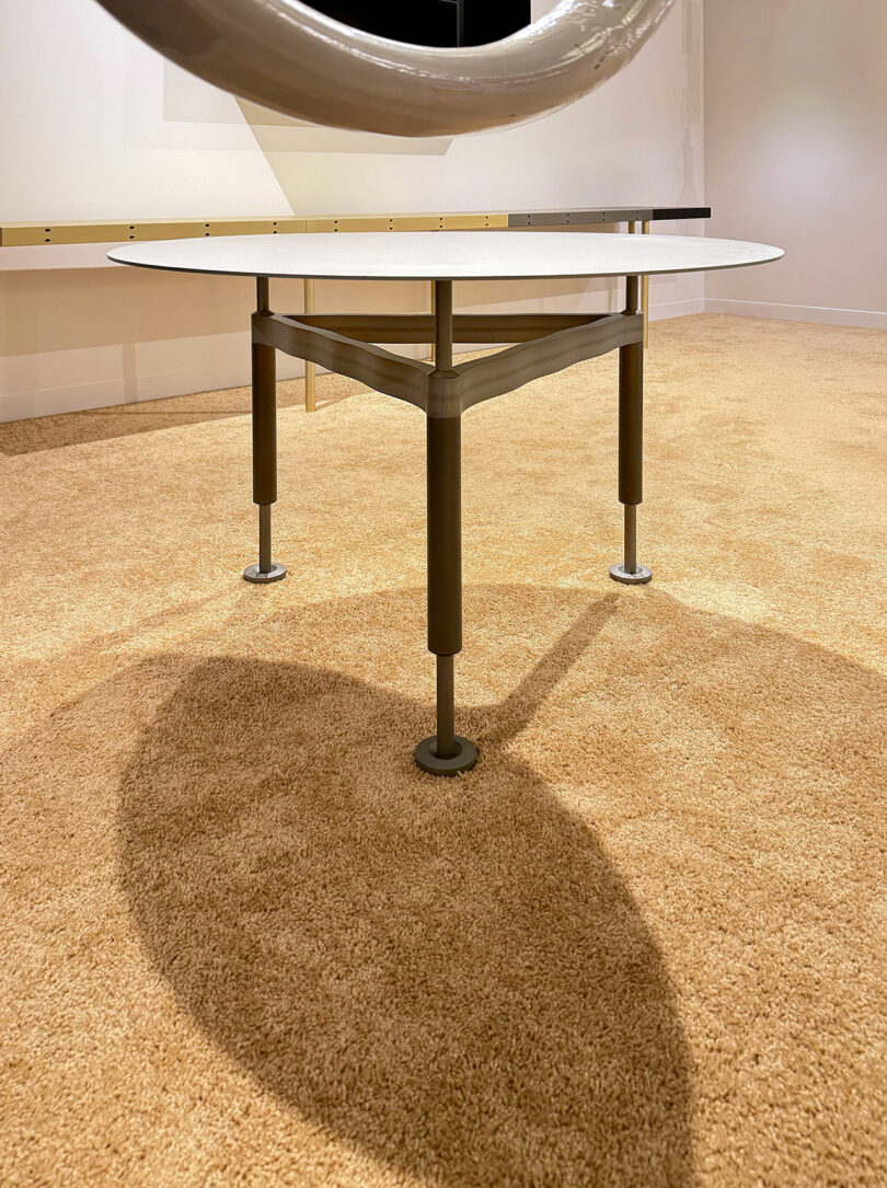 A round table with a white top and three dark metal legs stands on light brown carpet, casting a shadow. A partial circular object appears at the top of the image.